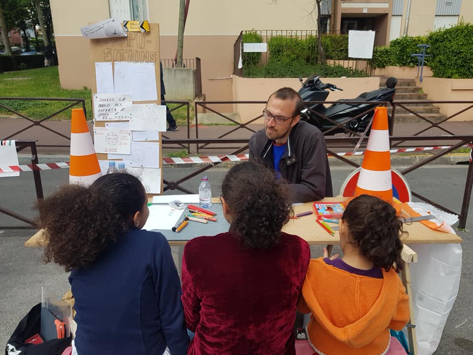 3 jeunes à une table d’atelier avec Jan Vormann, avec des plots orange sur la table, dans le cadre du chantier partagé « Déviations » de la Société du Grand Paris