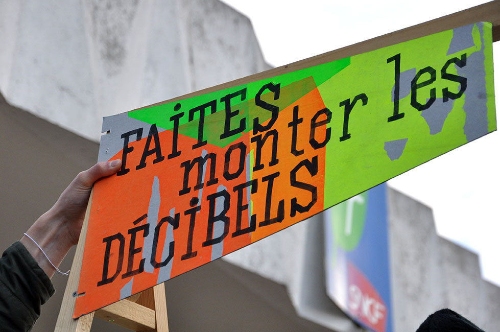 Drapeau en carton coloré sur lequel est inscrit « faites monter les décibels », aux abords du chantier des Ardoines dans le cadre du chantier partagé du collectif « Ne Rougissez Pas »