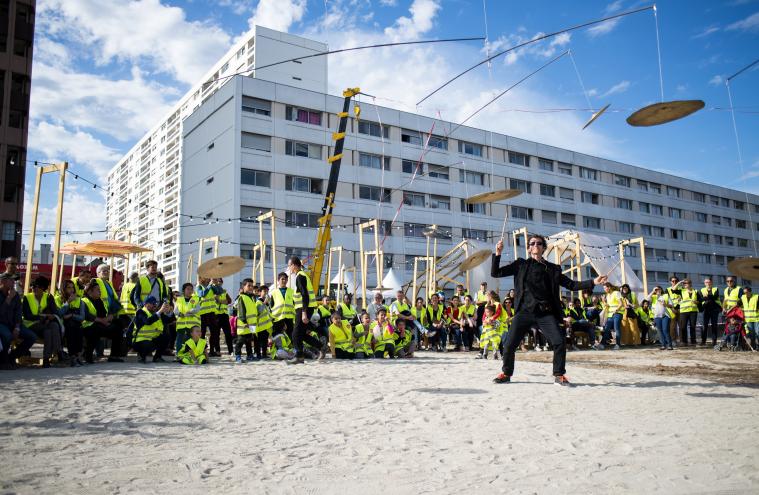 Performance de l’acrobate Cyril Hernandez avec le public en gilet jaune de chantier autour, de jour, lors du KM4 à La Courneuve