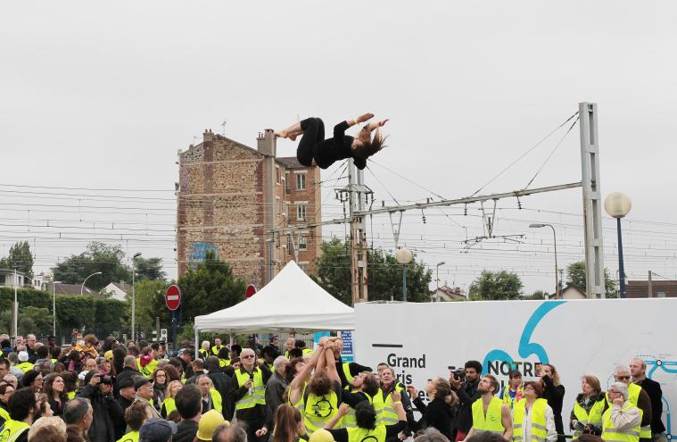 Performance d’acrobates lors du KM1 à Fort-d’Issy-Vanves-Clamart-Malakoff, avec le public et les acrobates en gilet jaune de chantier, de jour, devant un café 