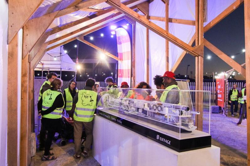 Maquette de tunnelier dans une boîte de verre avec du public en gilet jaune de chantier, lors du KM4 à La Courneuve